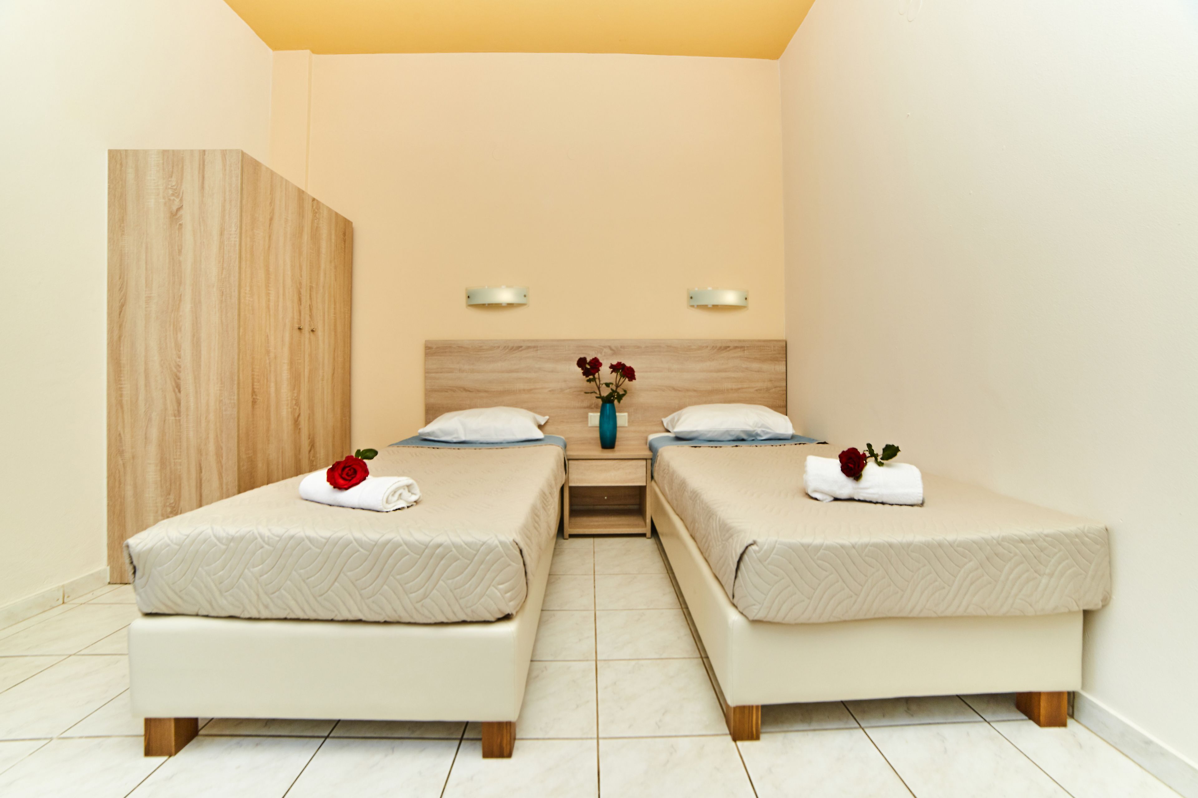 A hotel room with two beds, a wooden wardrobe, a nightstand with a vase of flowers, and a wall-mounted light fixture.