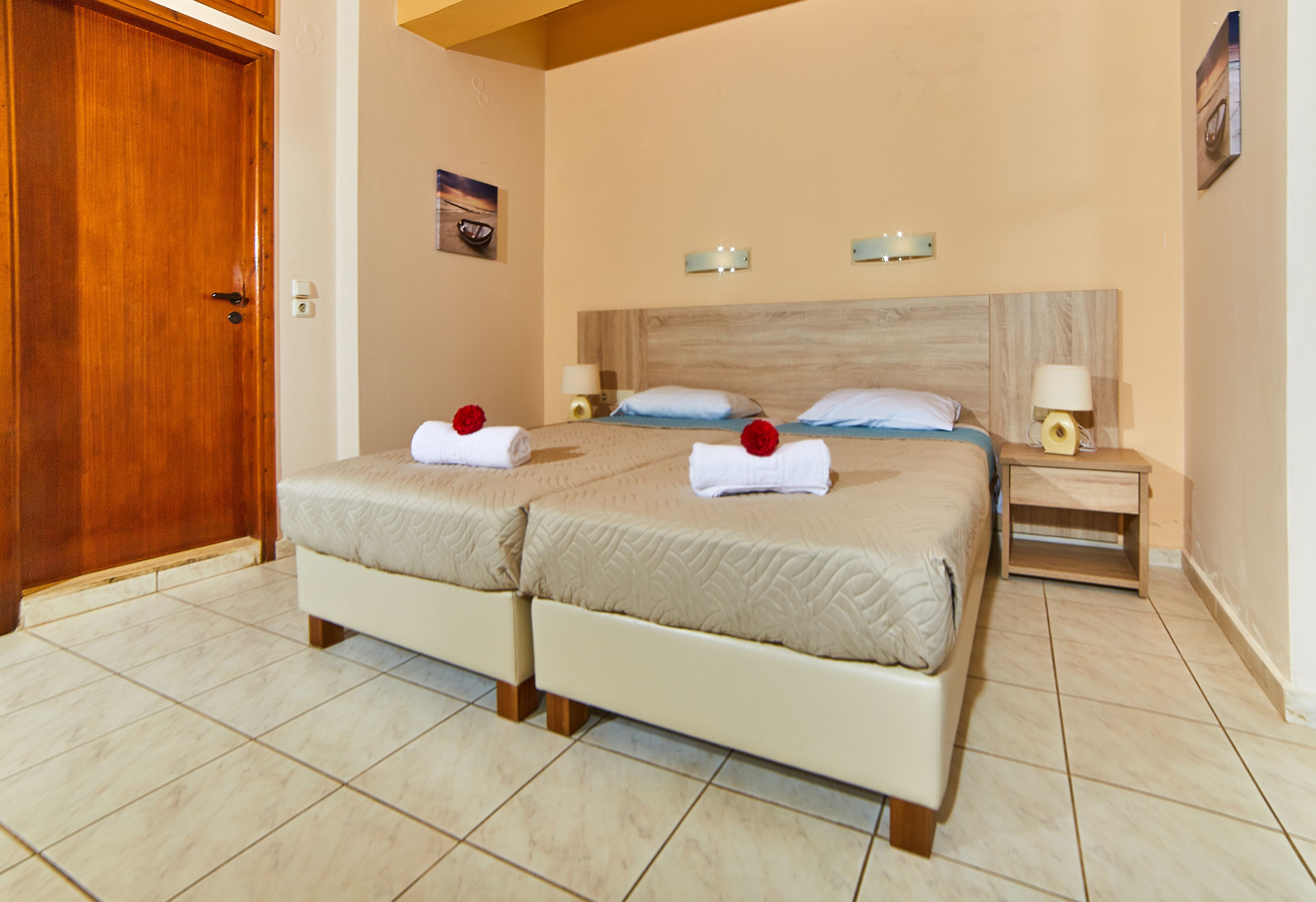 A hotel room with two beds, a wooden door, a wall-mounted light fixture, and a small bedside table with a lamp.