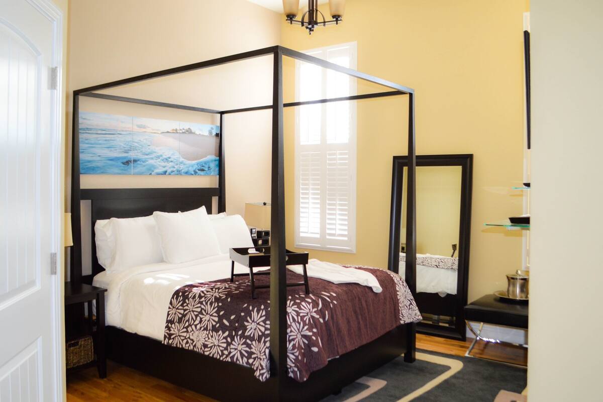 A four-poster bed with a dark frame, white bedding, and a floral-patterned throw. A painting of a beach scene is above the bed.