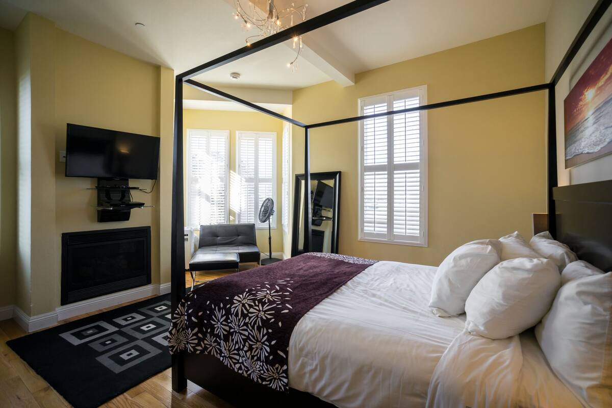 A bedroom with a four-poster bed, a flat-screen TV mounted on the wall, a fireplace, and a large window with blinds.