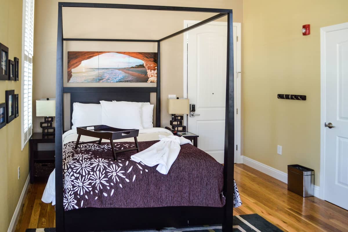 A four-poster bed with a floral bedspread, a painting above the bed, a nightstand with a lamp, and a door with a red emergency light.