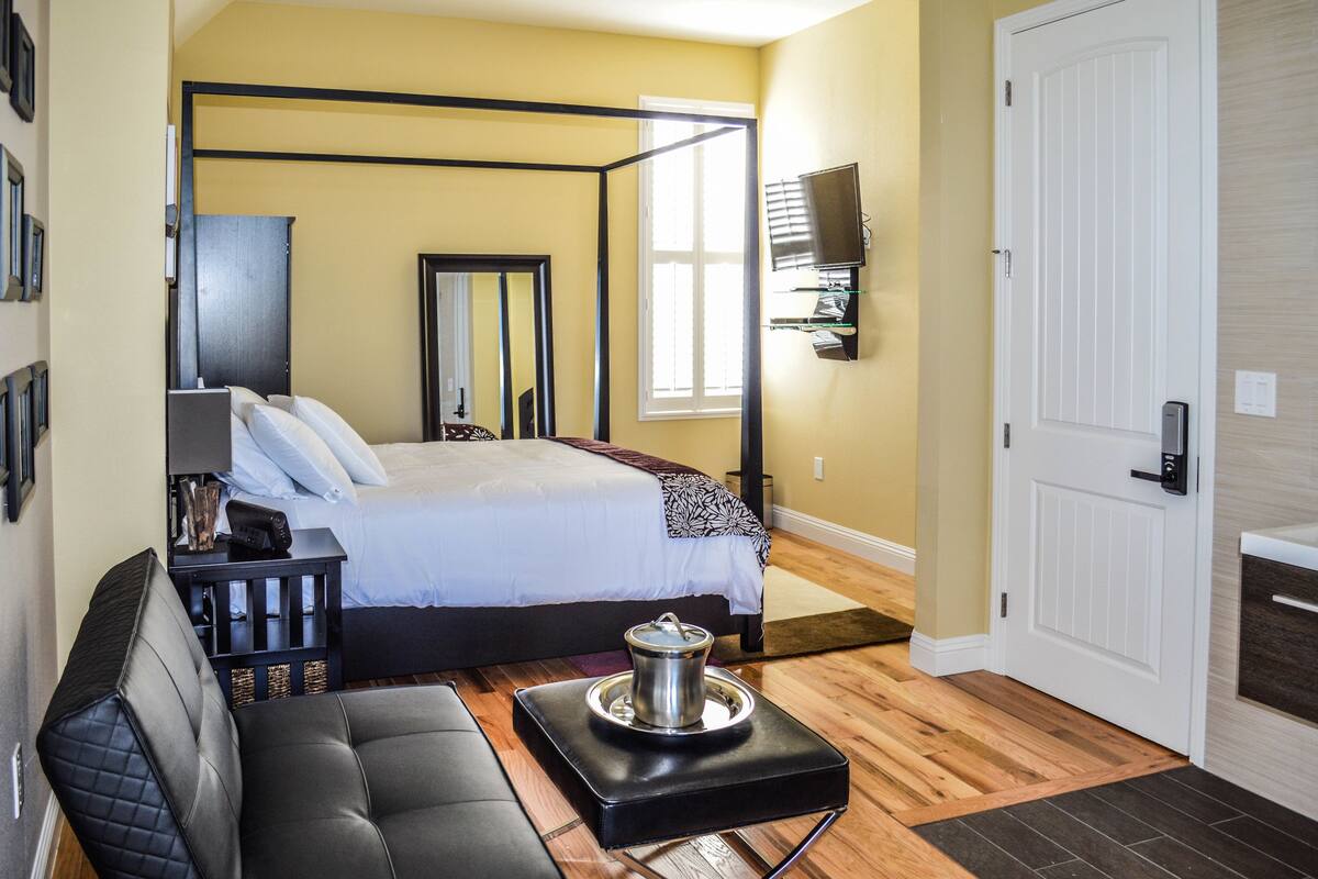 A bedroom with a four-poster bed, a black leather sofa, a bedside table with a lamp, and a TV mounted on the wall.