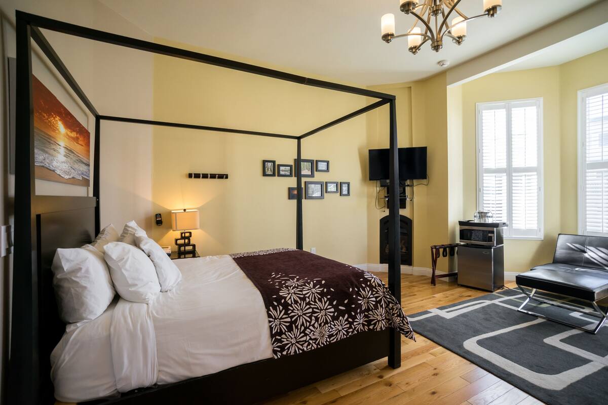A four-poster bed with white linens and a patterned throw, a flat-screen TV, a fireplace, and a leather sofa in a well-lit room with wooden flooring.