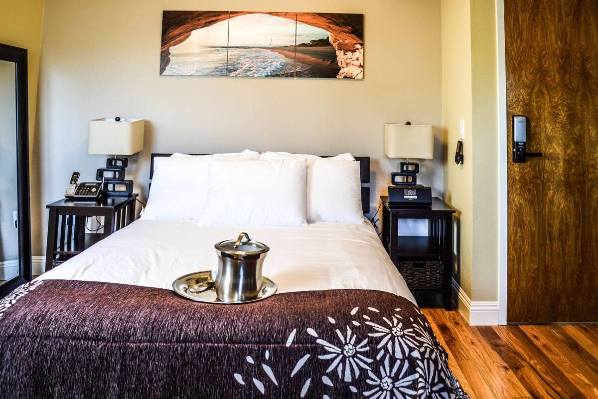 A neatly made bed with a tray of tea, two bedside tables with lamps, a painting of a beach scene, and a wooden door.