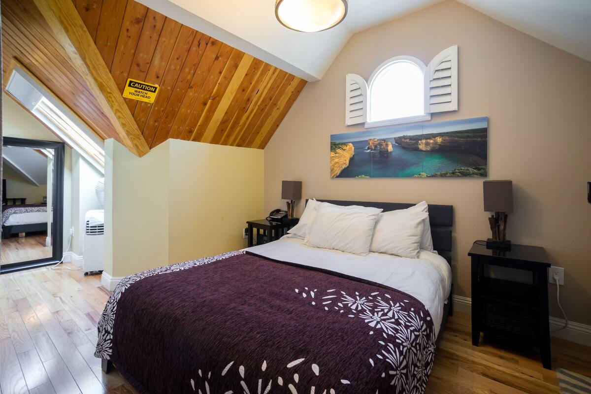 A bedroom with a wooden ceiling, a bed with a white and purple bedspread, a painting of a coastal scene, and a caution sign.