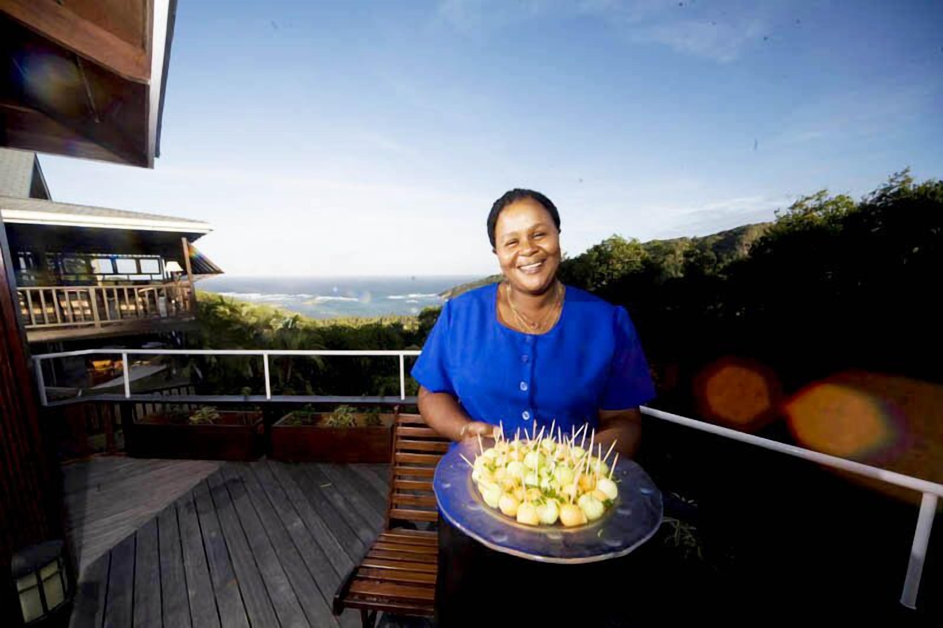 Una persona sostiene una bandeja con comida en una terraza de madera, con vista al océano y colinas al fondo.