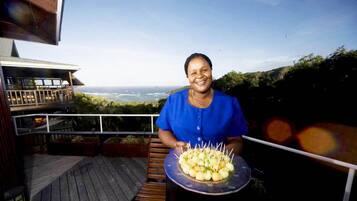 Una persona sostiene una bandeja con comida en una terraza de madera, con vista al océano y colinas al fondo.
