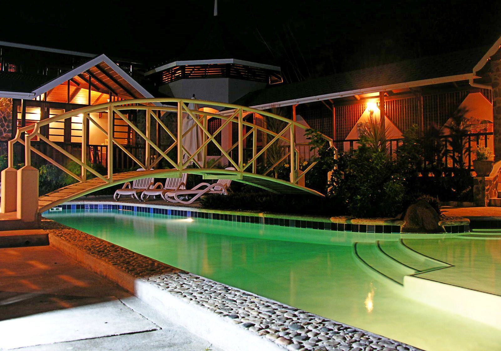 Ein Schwimmbad mit einer Brücke, Liegestühlen und einem Gebäude mit warmer Beleuchtung.