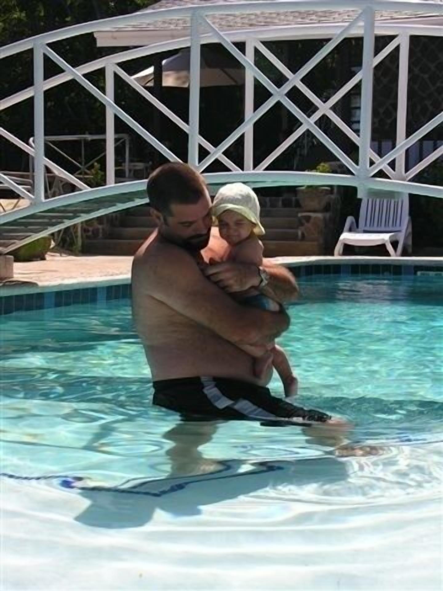Un hombre sostiene a un niño en una piscina, con una estructura de quiosco blanco al fondo.