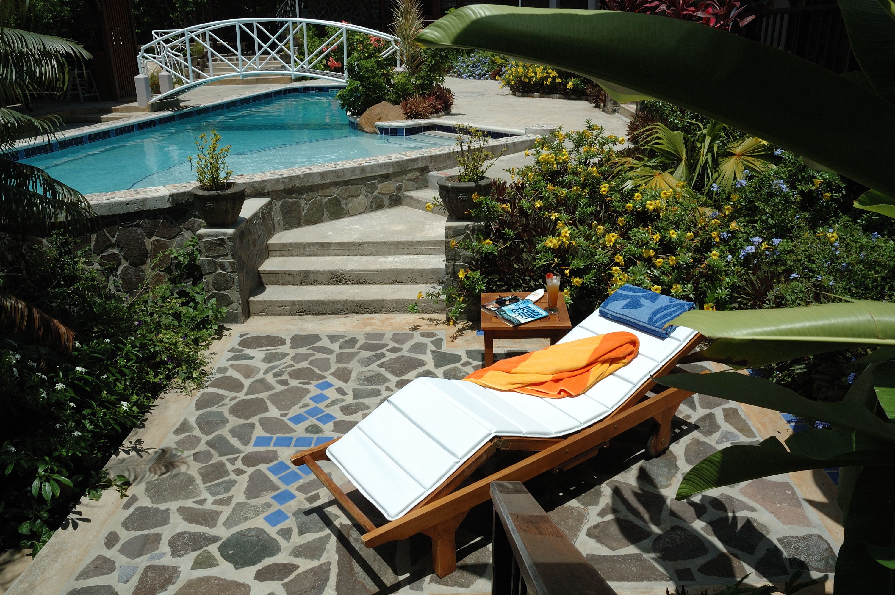 A pool area with a lounge chair, a wooden bridge, and a stone-tiled deck.