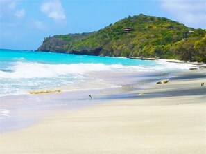 A beach with clear turquoise water, a sandy shore, and a hill with vegetation and a structure on top.