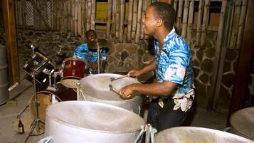 A person playing a drum set in a rustic setting with stone walls and wooden structures.