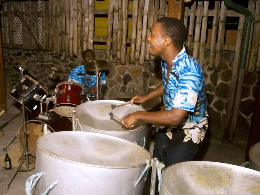 A person playing a drum set in a rustic setting with stone walls and wooden structures.