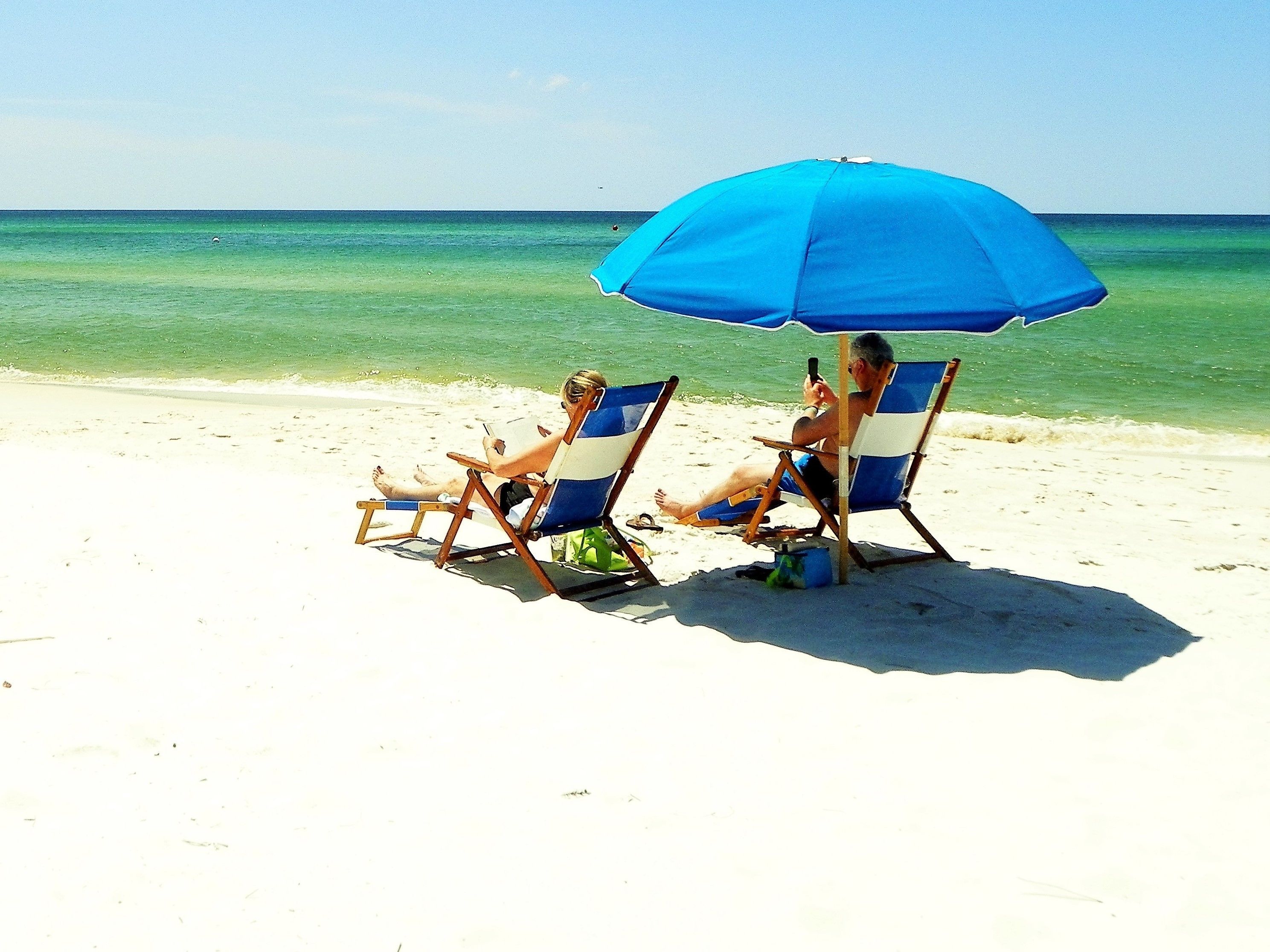 Duas pessoas sentadas em cadeiras de praia sob um guarda-sol azul numa praia de areia com águas cristalinas.
