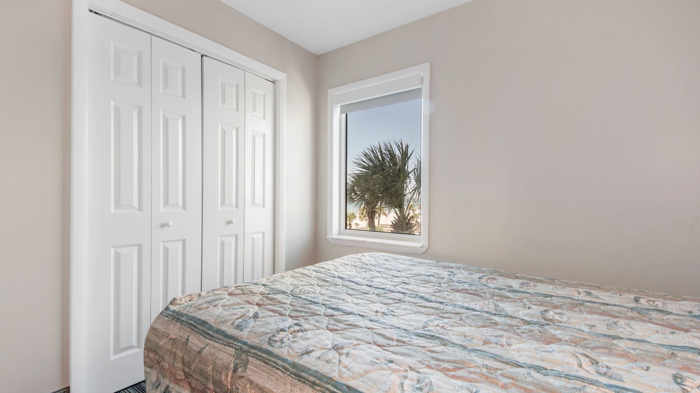 A bedroom with a bed, a window with a view of palm trees, and a white wardrobe.