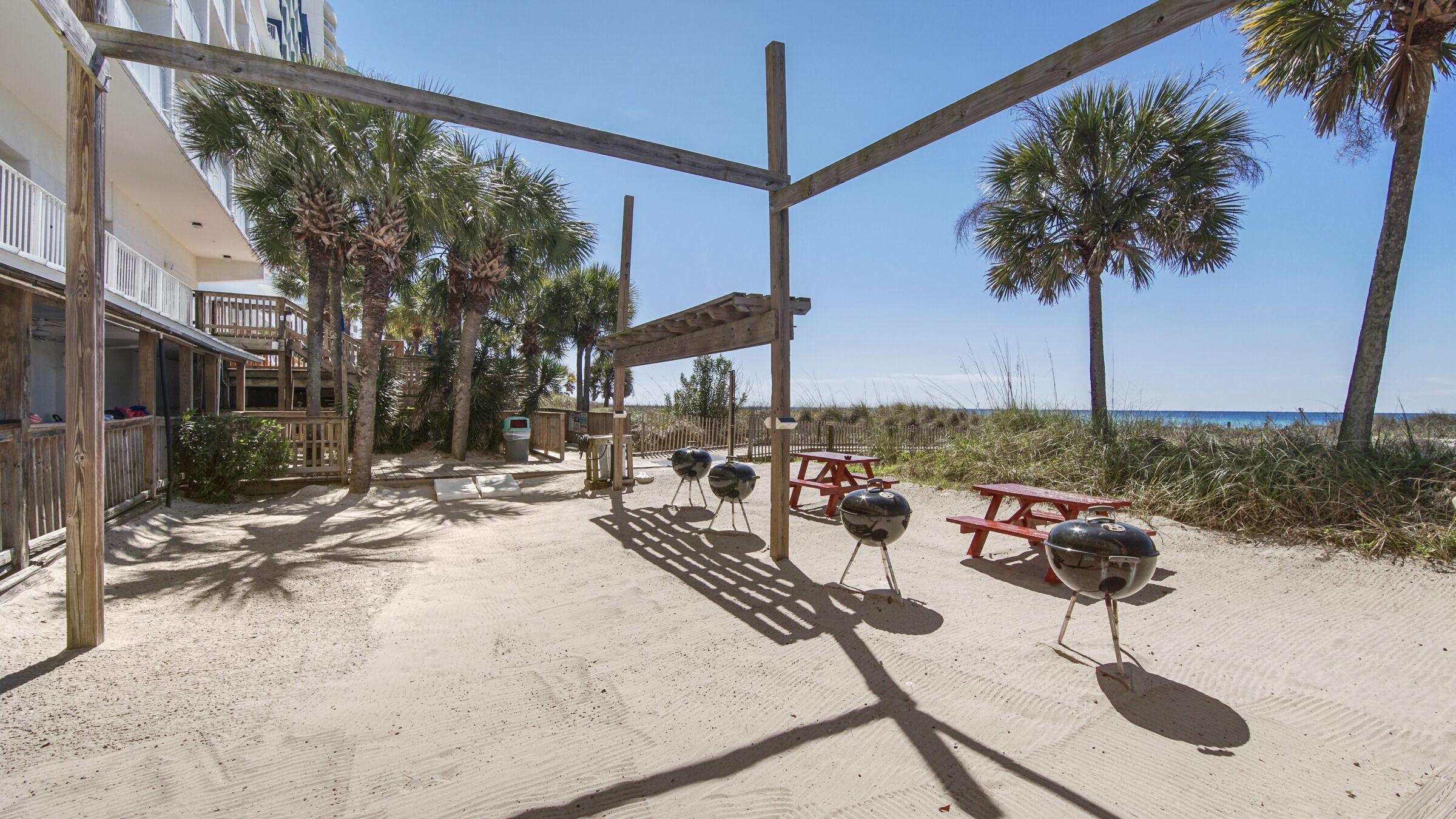 A beachside patio with wooden pergolas, red picnic tables, and barbecue grills.