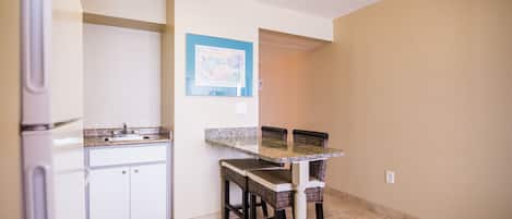 Cocina compacta con gabinetes blancos, encimera de granito y comedor con sillas de mimbre.