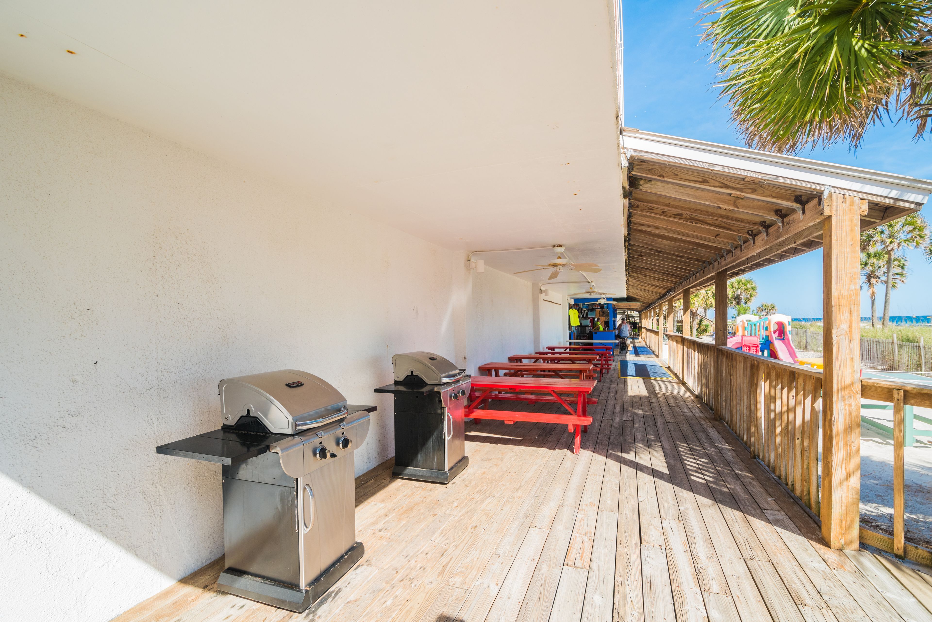 A covered outdoor area with barbecue grills, wooden benches, and a view of the beach.