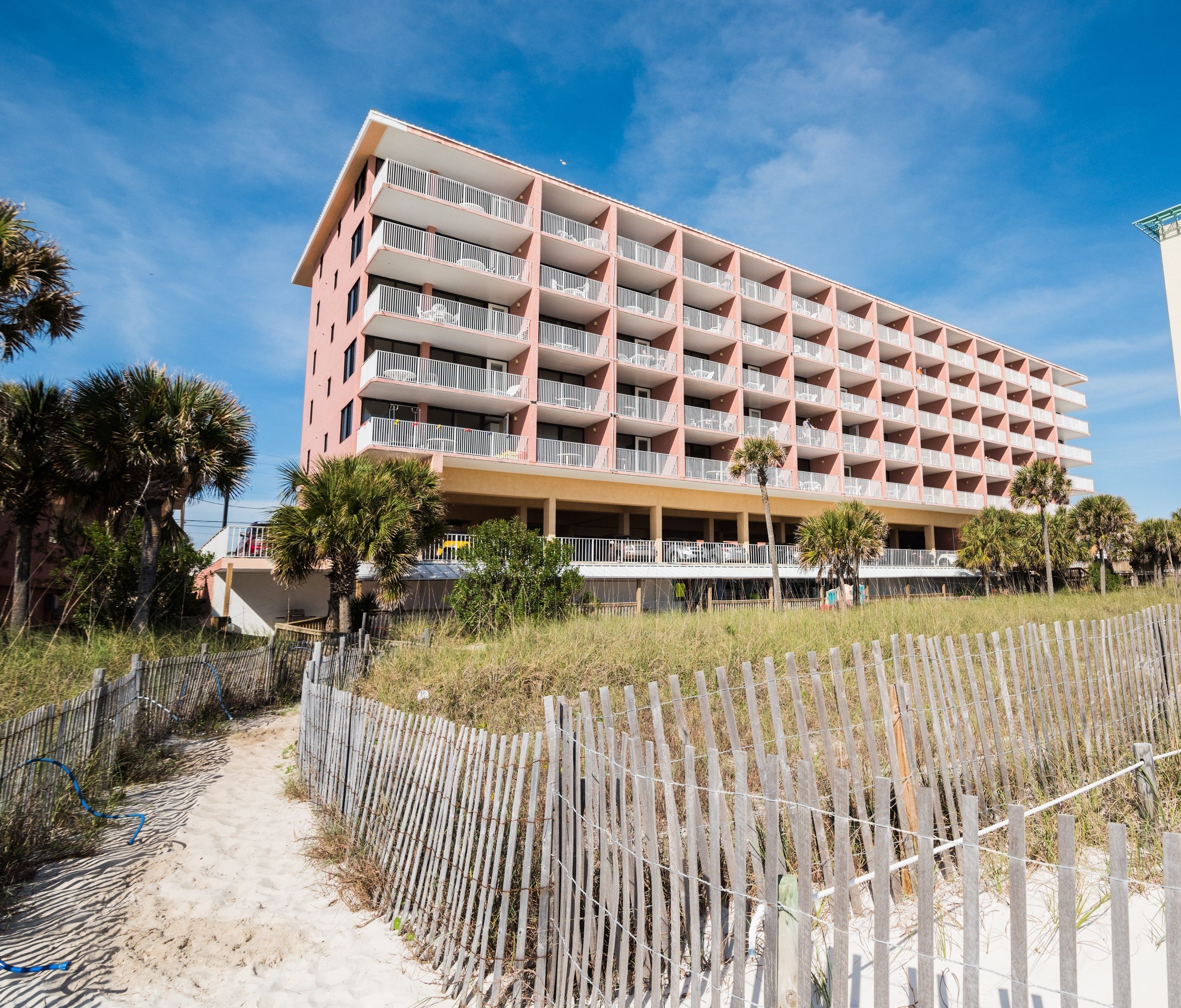 A multi-story hotel with balconies, a sandy path leading to the entrance, and a wooden fence.
