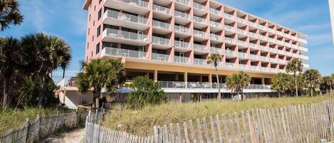 A multi-story hotel with balconies, a sandy path leading to the entrance, and a wooden fence.