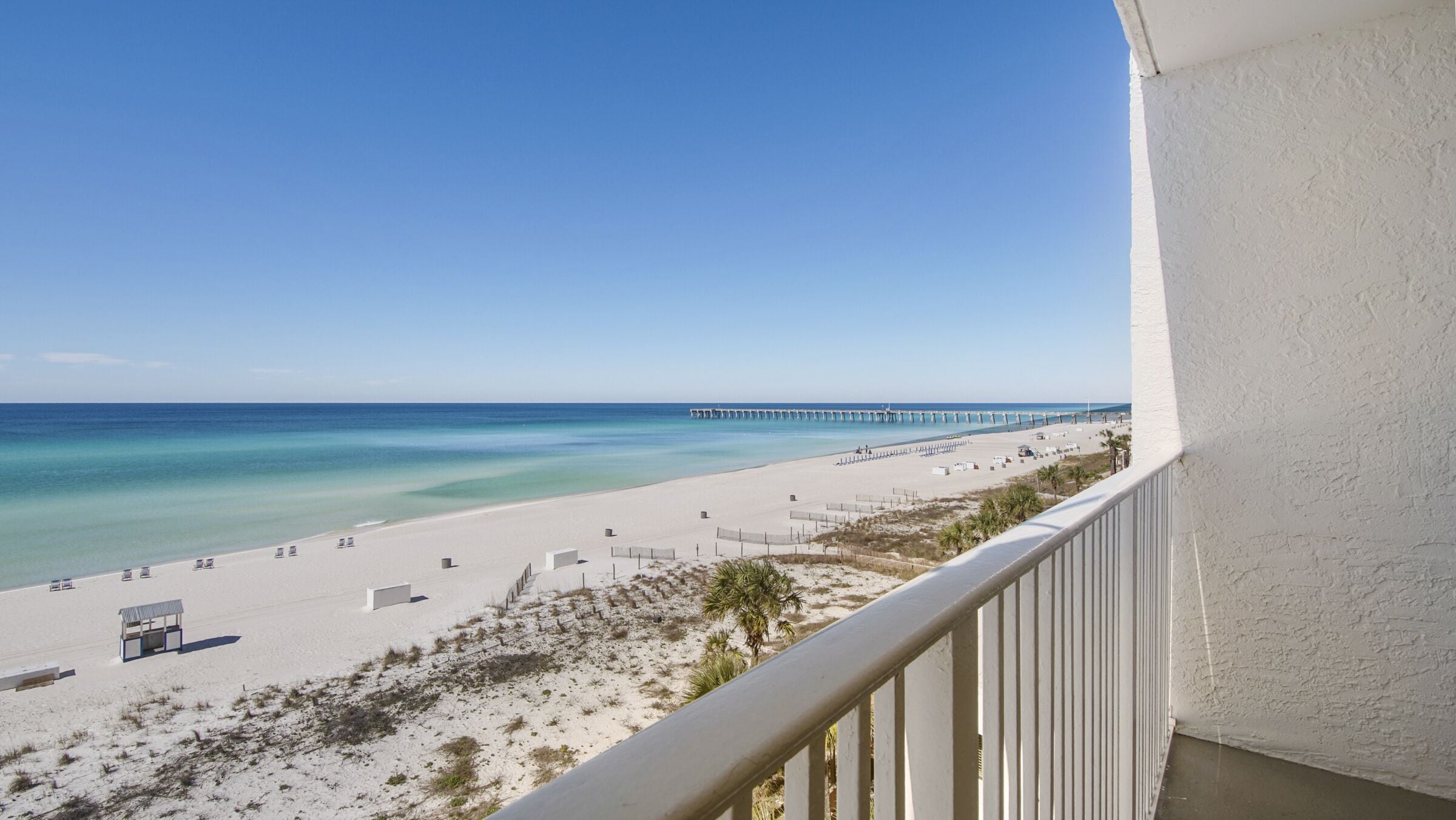 A beach view from a balcony with a clear sky, a sandy beach, and a pier in the distance.