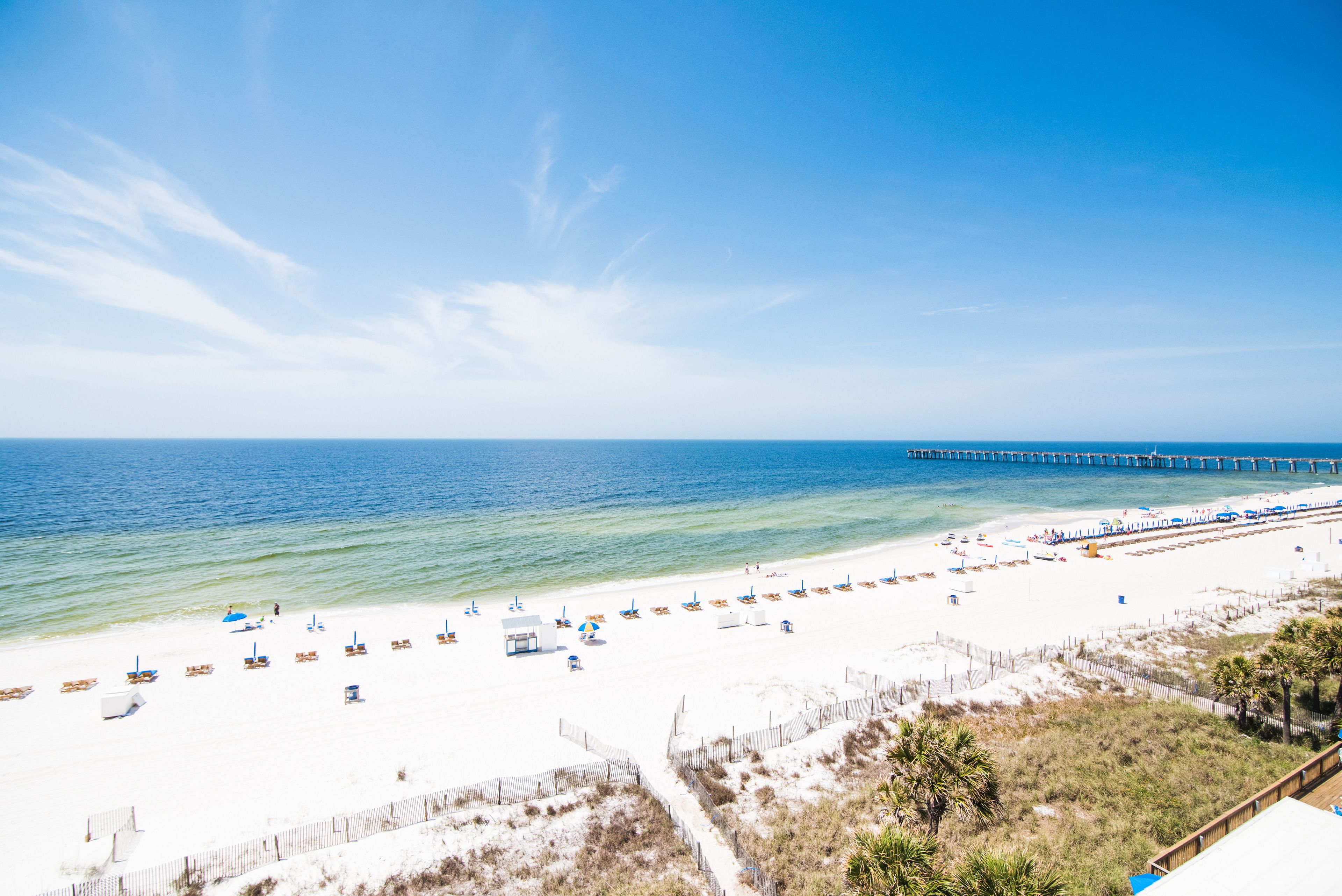 A beach with white sand, a pier extending into the water, and beach umbrellas.