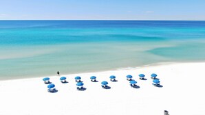 Une plage de sable blanc bordée d’une eau turquoise et limpide. Des parasols bleus et des chaises longues sont disposés sur le sable.