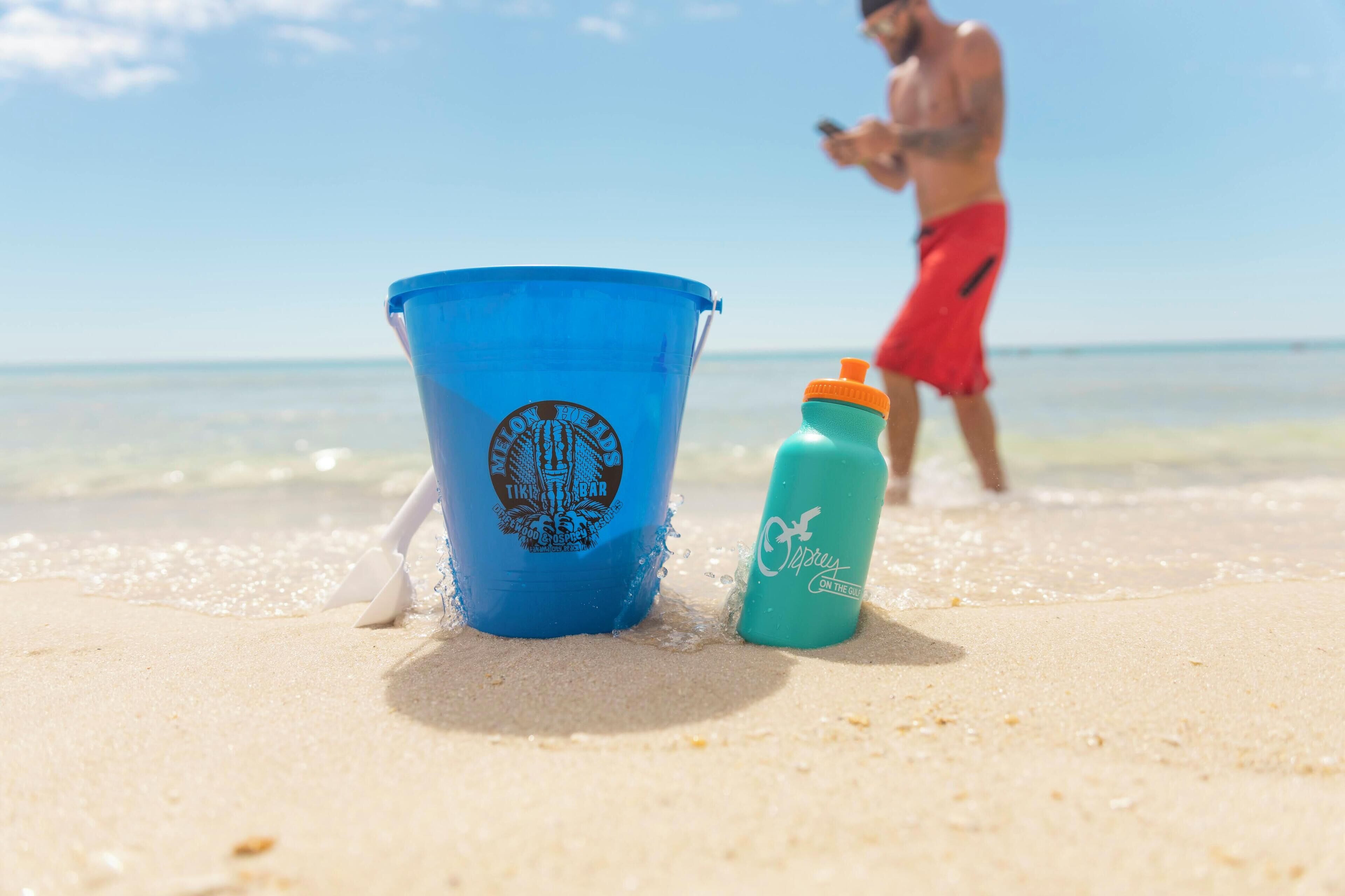 Un cubo azul y una botella de agua sobre una playa arenosa, con una persona al fondo.
