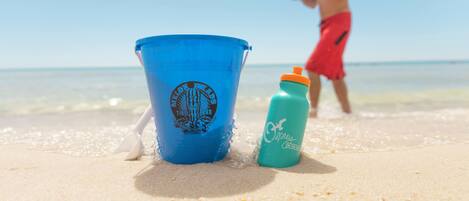 Un cubo azul y una botella de agua sobre una playa arenosa, con una persona al fondo.