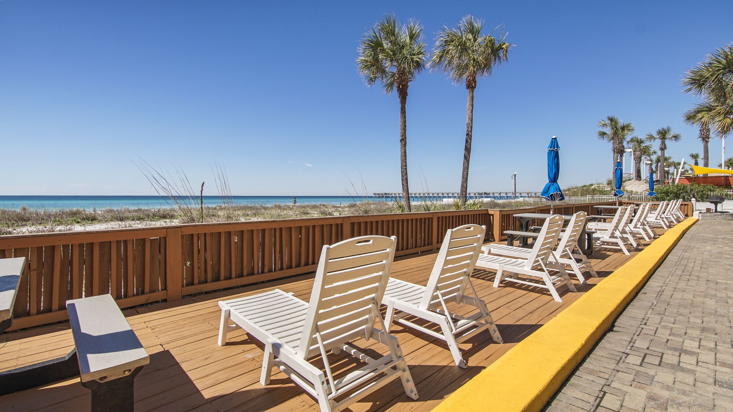 A wooden deck with white lounge chairs and a wooden table, overlooking a beach with palm trees and a pier.