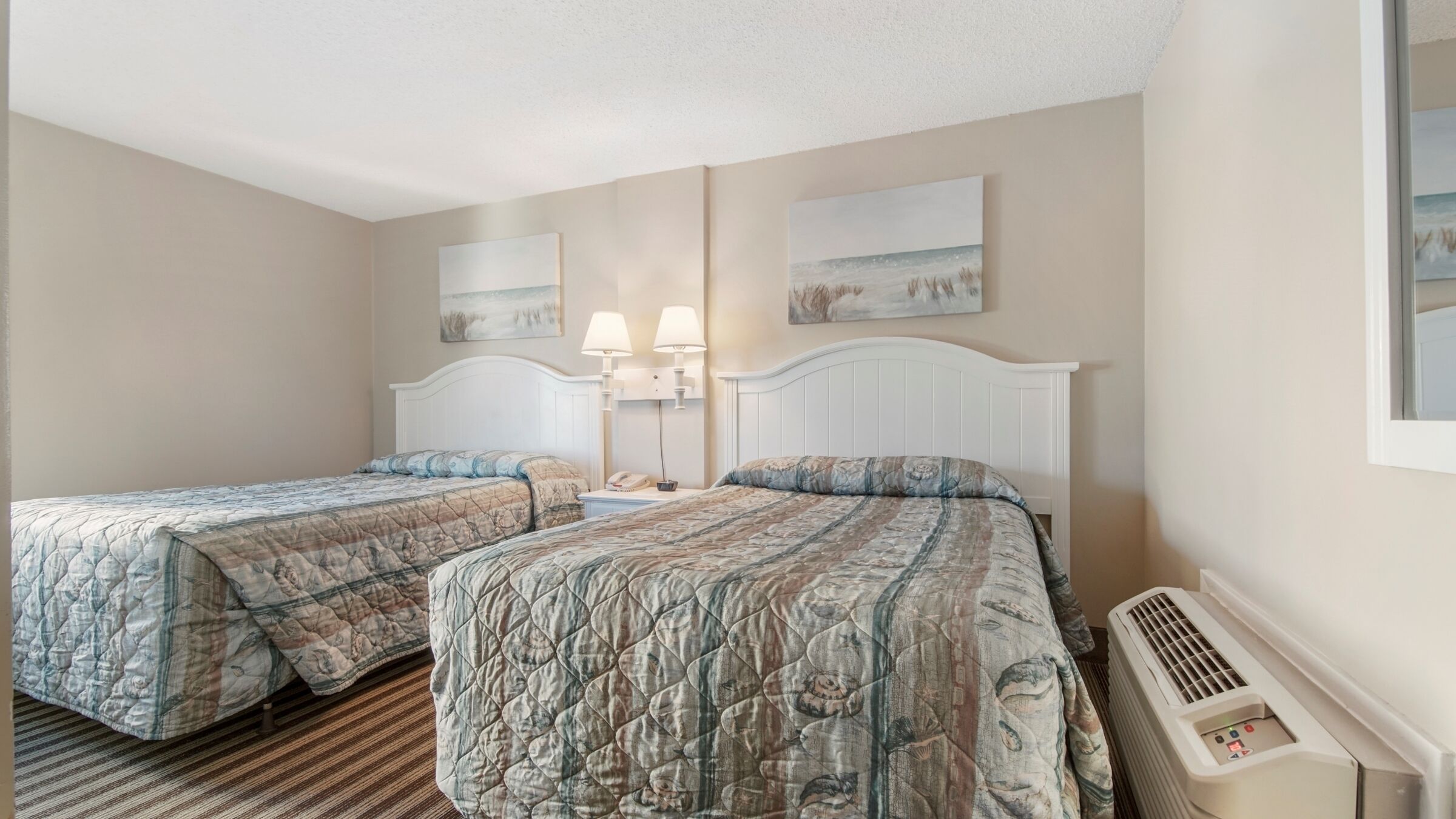 A hotel room with two beds, a white headboard, a bedside table with a lamp, and a wall-mounted air conditioning unit.