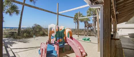 A beachside playground with a slide and climbing structures, surrounded by palm trees and a clear sky.