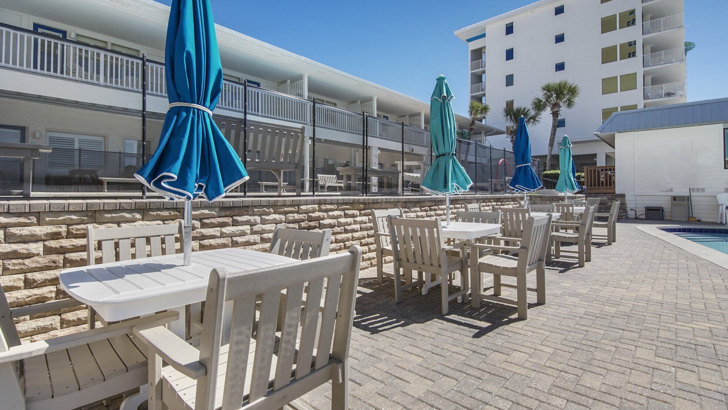 An outdoor seating area with white chairs and tables, blue umbrellas, and a pool in the background.