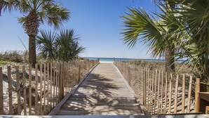 Une promenade en bois menant à une plage, bordée de palmiers et d’un sentier sablonneux.