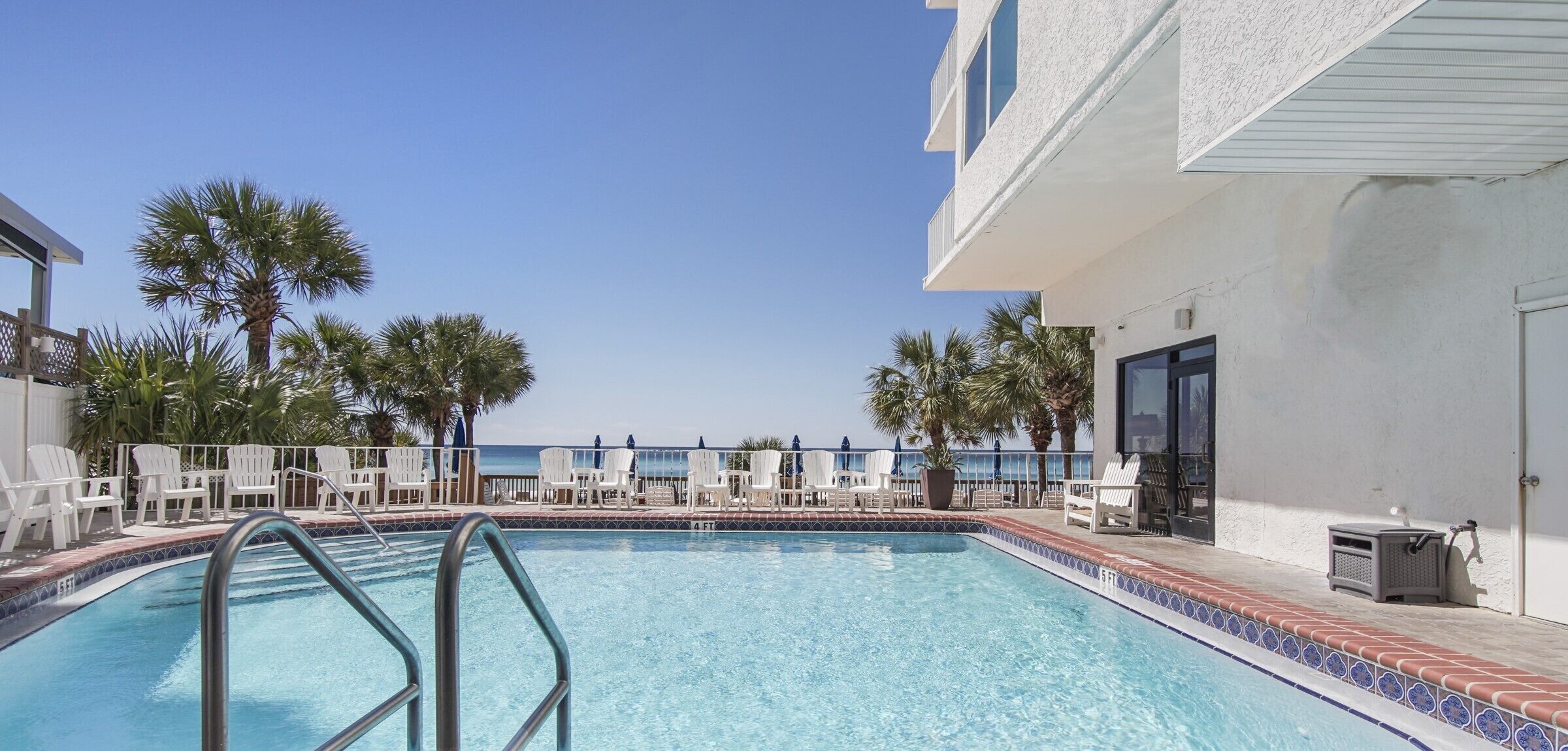 A swimming pool with white chairs and palm trees, a clear sky, and a view of the ocean.