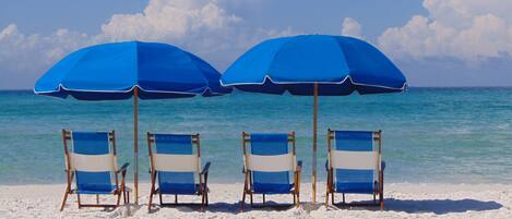 Four beach chairs under blue umbrellas on a sandy beach with clear blue water and a cloudy sky.