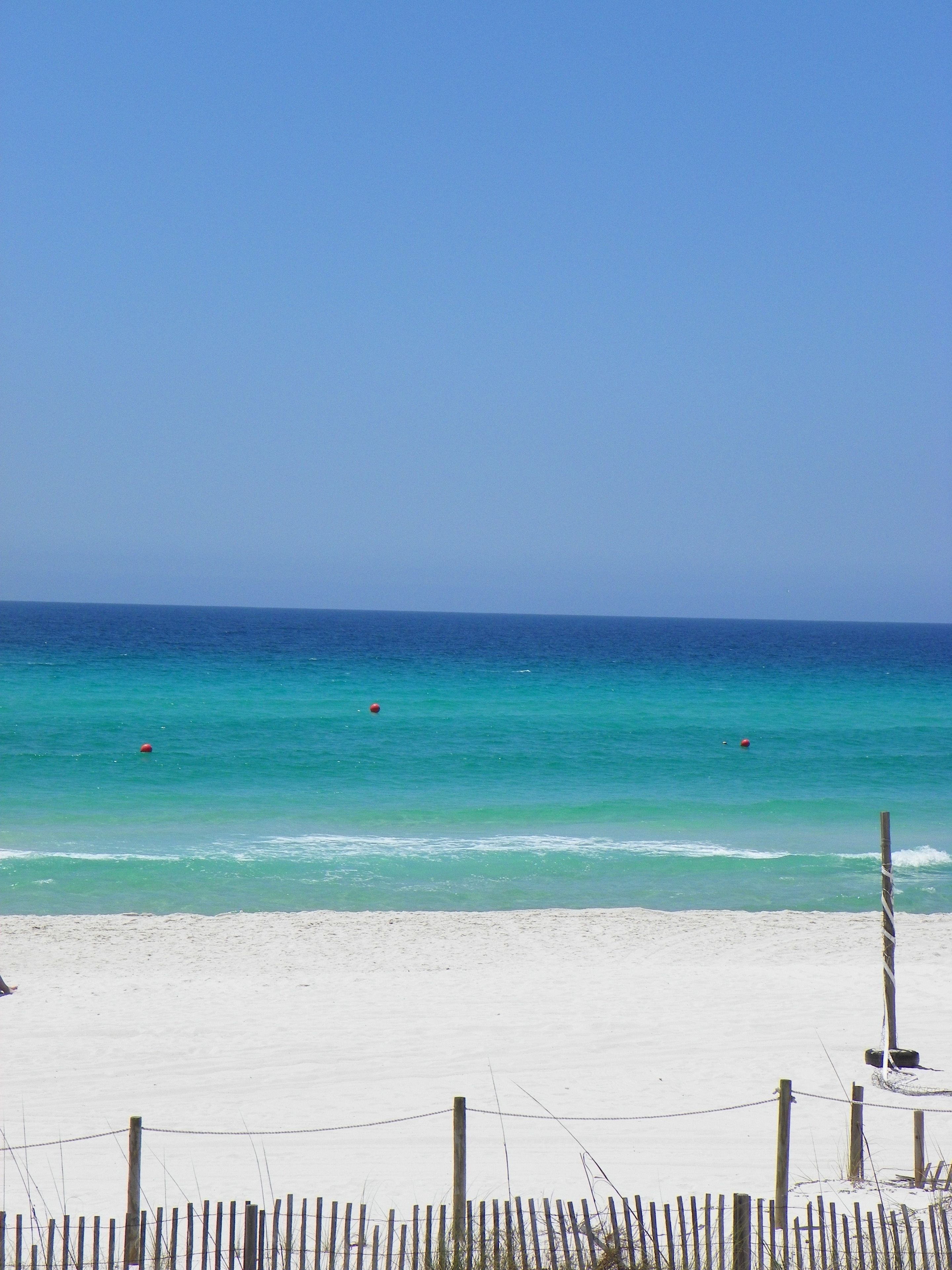 A beach with clear turquoise water, a sandy shore, and a wooden fence in the foreground.
