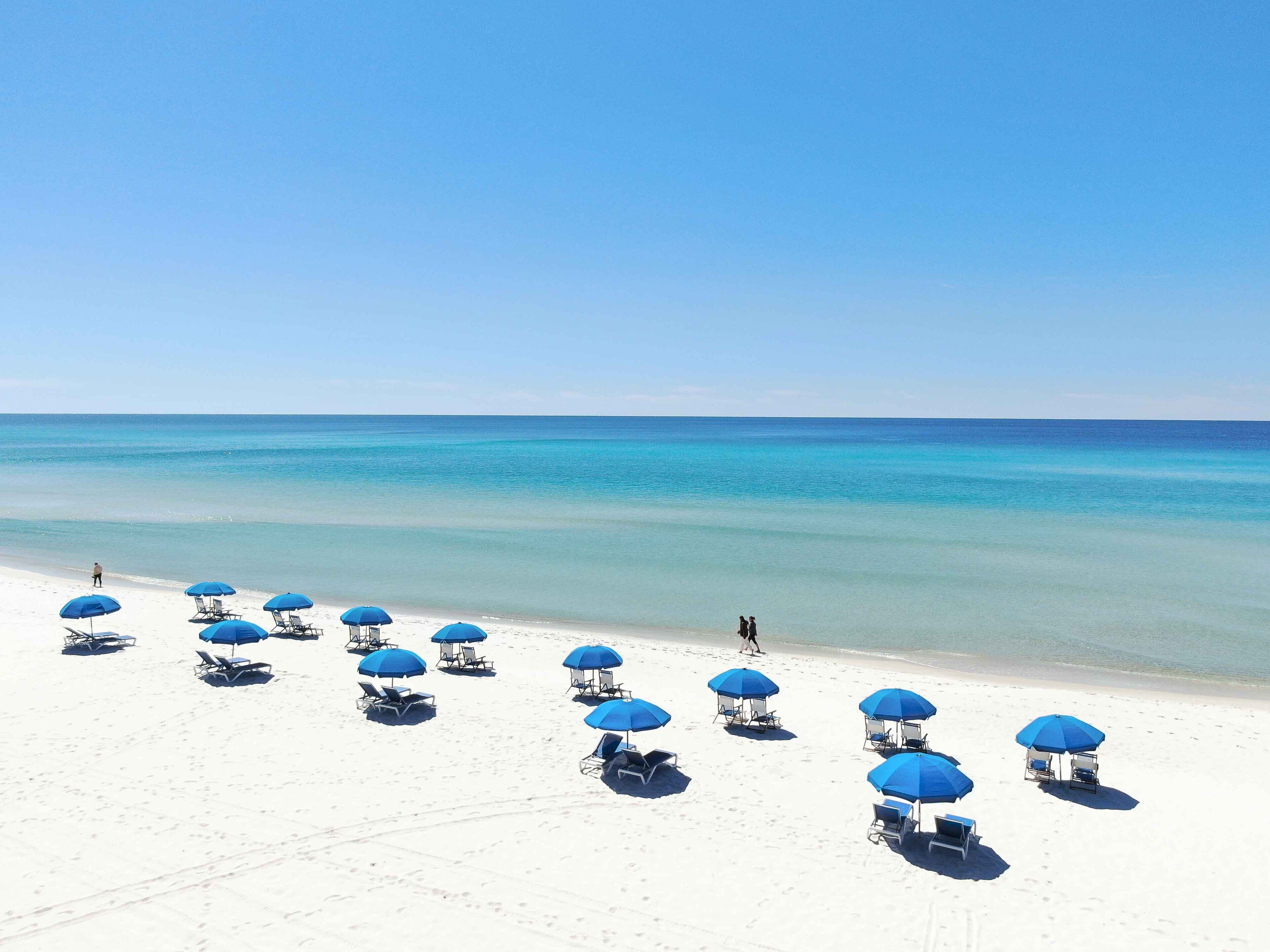 Uma praia com areia branca e água azul cristalina. Vários guarda-sóis azuis e cadeiras de praia estão dispostos ao longo da costa.