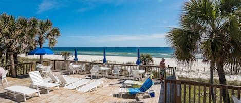 A wooden deck with white lounge chairs and blue umbrellas overlooking a beach.