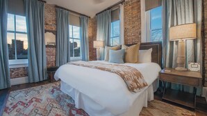Habitación de hotel con una cama grande, dos mesitas de noche con lámparas, un tapete estampado y una pared de ladrillo.