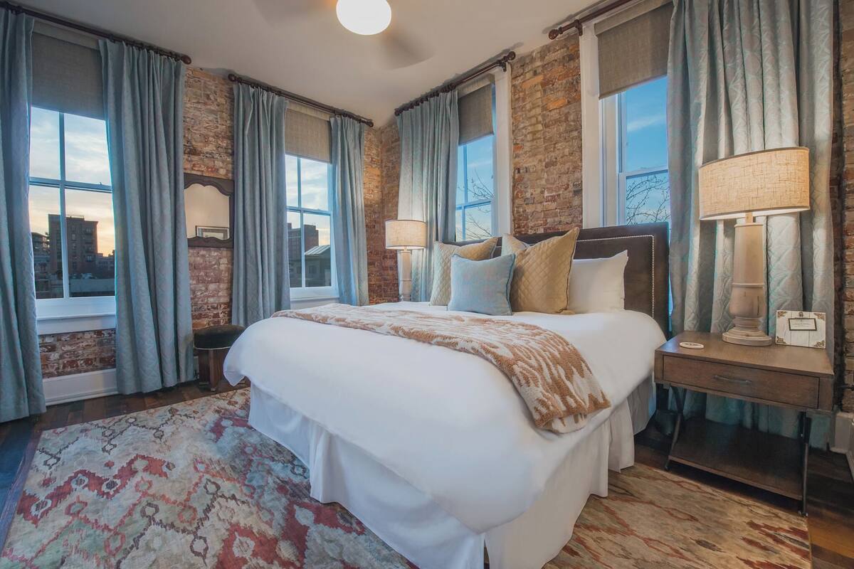 Habitación de hotel con una cama grande, dos mesitas de noche con lámparas, un tapete estampado y una pared de ladrillo.