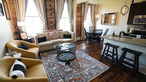 Una sala de estar con un sofá, sillones, una mesa de centro y un tapete estampado. Hay un comedor con una mesa y sillas, y una cocina con microondas y armarios.