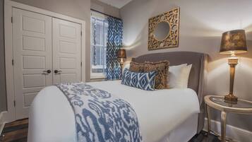 A bedroom with a large bed, a bedside table with a lamp, a window with patterned curtains, and a decorative mirror on the wall.