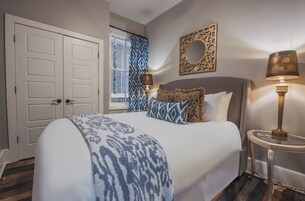A bedroom with a large bed, a bedside table with a lamp, a window with patterned curtains, and a decorative mirror on the wall.
