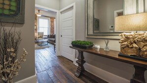 Un pasillo con piso de madera, una puerta blanca, un espejo, una lámpara y una mesita con una planta y una vela.