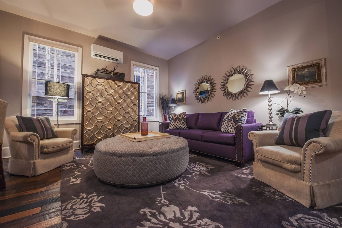 Una sala de estar con un otoman redondo, dos sillones, un sofá morado y espejos decorativos en la pared.