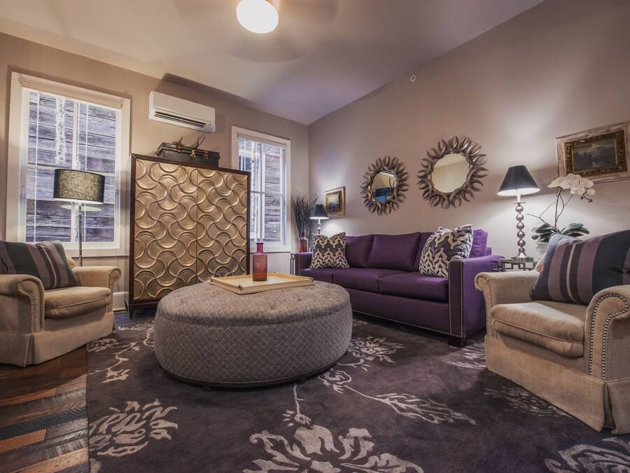 A living room with a round ottoman, two armchairs, a purple sofa, and decorative mirrors on the wall.