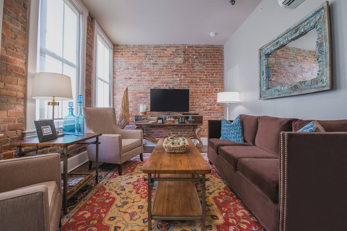 Una sala de estar con pared de ladrillo, televisor, mesa de centro de madera y alfombra estampada.