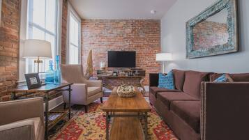 Una sala de estar con pared de ladrillo, televisor, mesa de centro de madera y alfombra estampada.