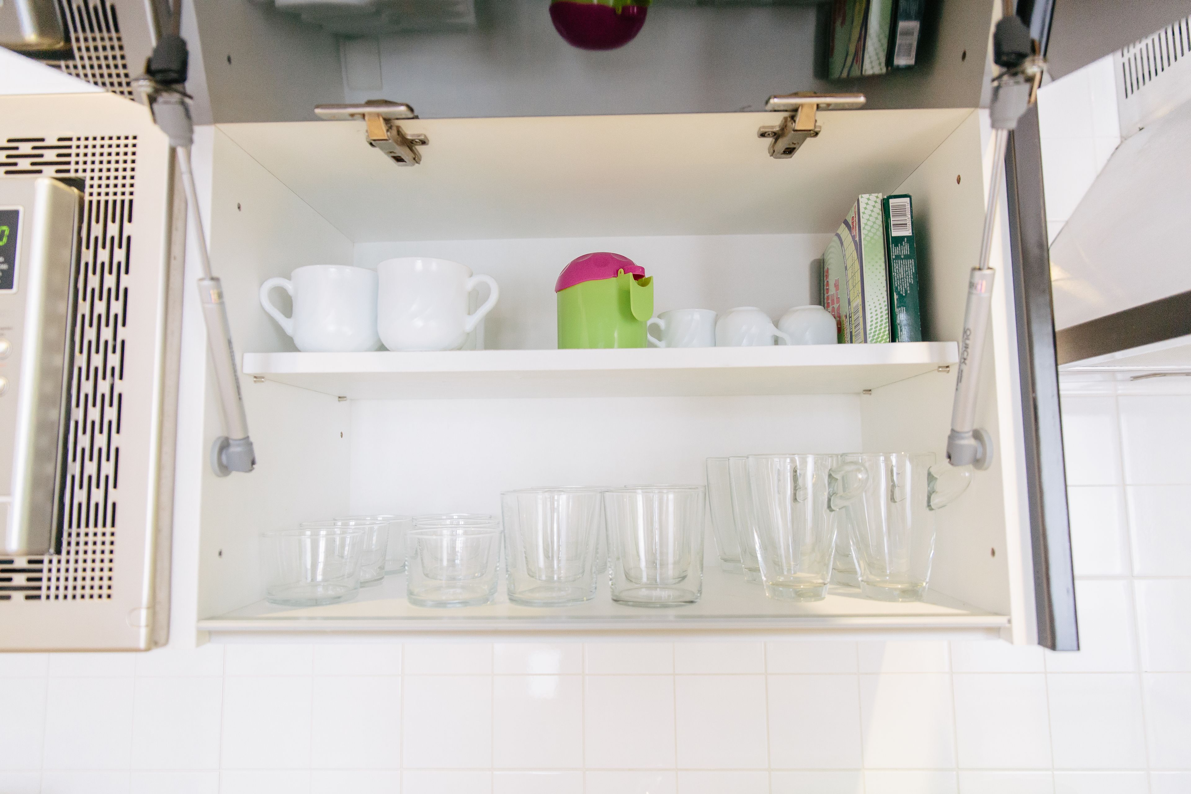 A kitchen with white cabinets, a shelf with white cups and a green mug, and a white tiled backsplash.